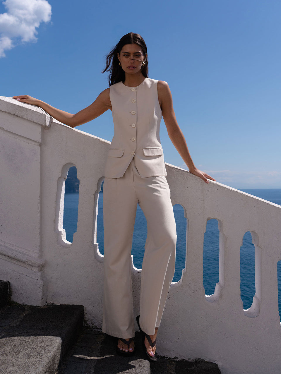 Woman in a beige suit set standing on a balcony with a blue sky and ocean view.