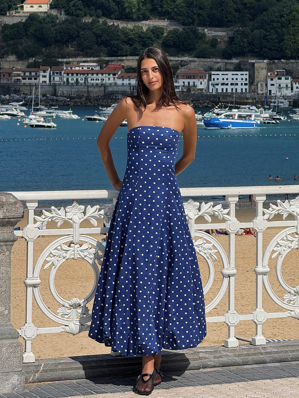 Woman in a blue polka dot dress standing by a waterfront with boats and buildings in the background.