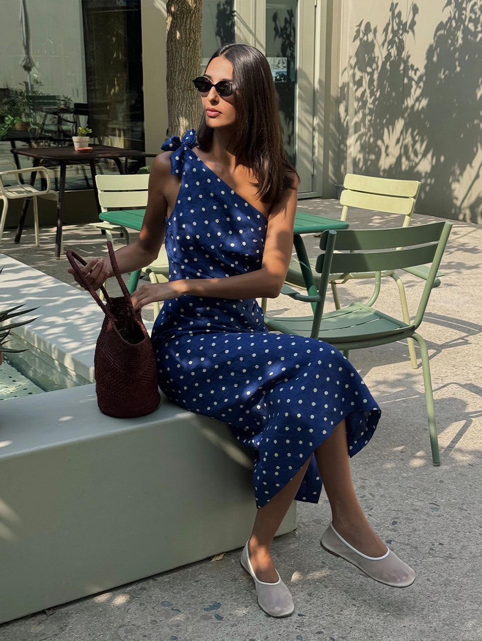 Woman in a blue polka dot dress sitting outdoors holding a brown handbag.