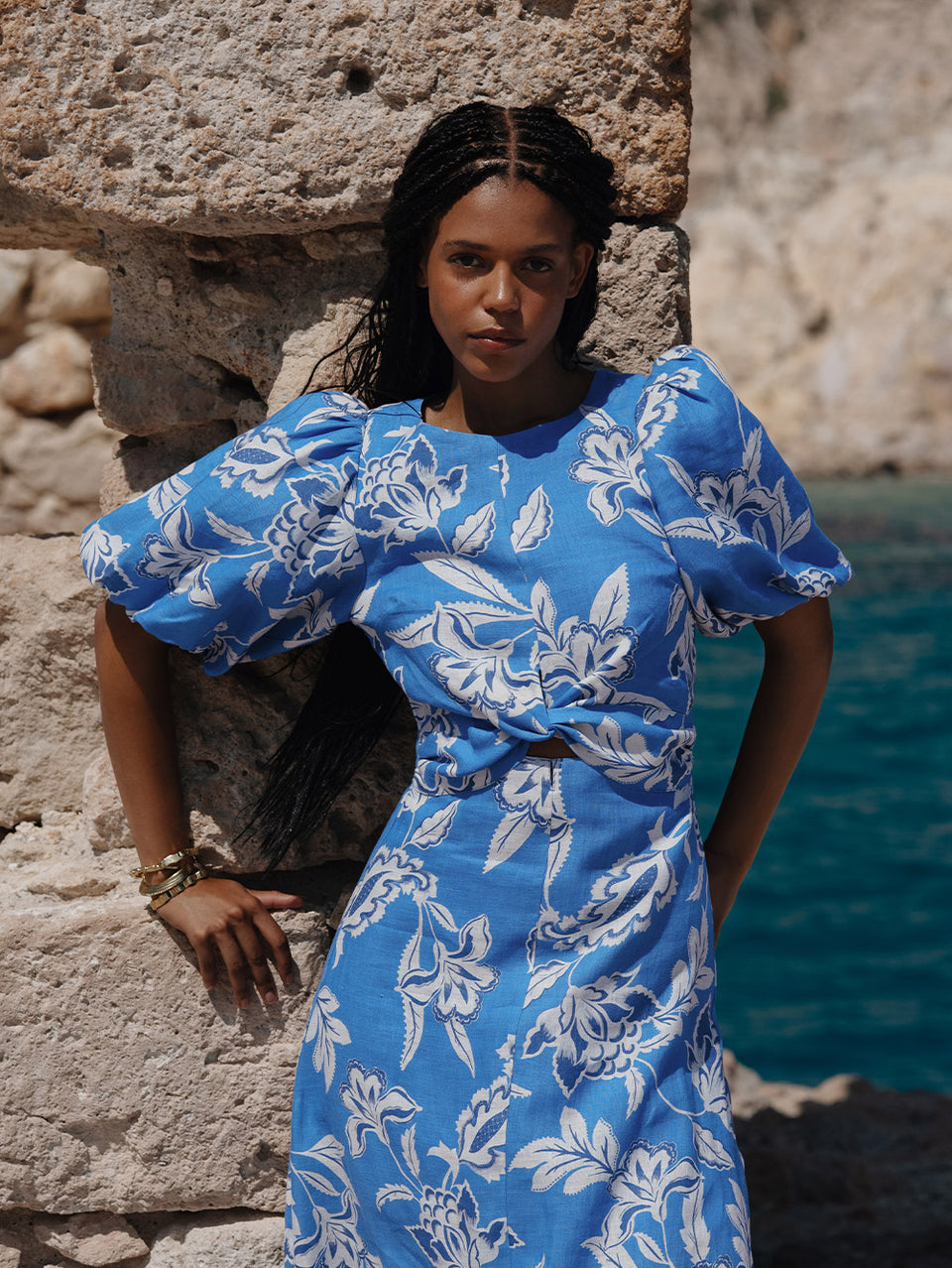 Woman in a blue floral dress standing against a stone wall with water in the background