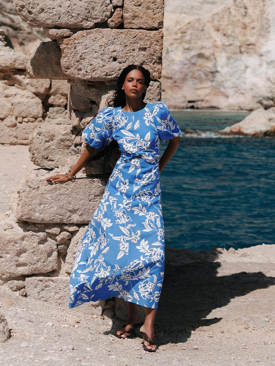 Woman in a blue floral dress standing against a stone wall by water.