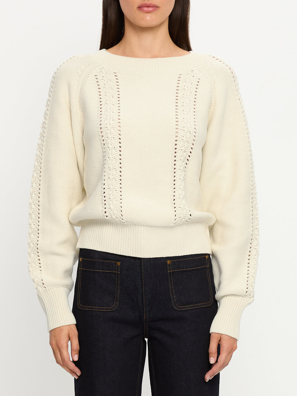 Person wearing a cream sweater with a textured pattern and dark jeans on a white background