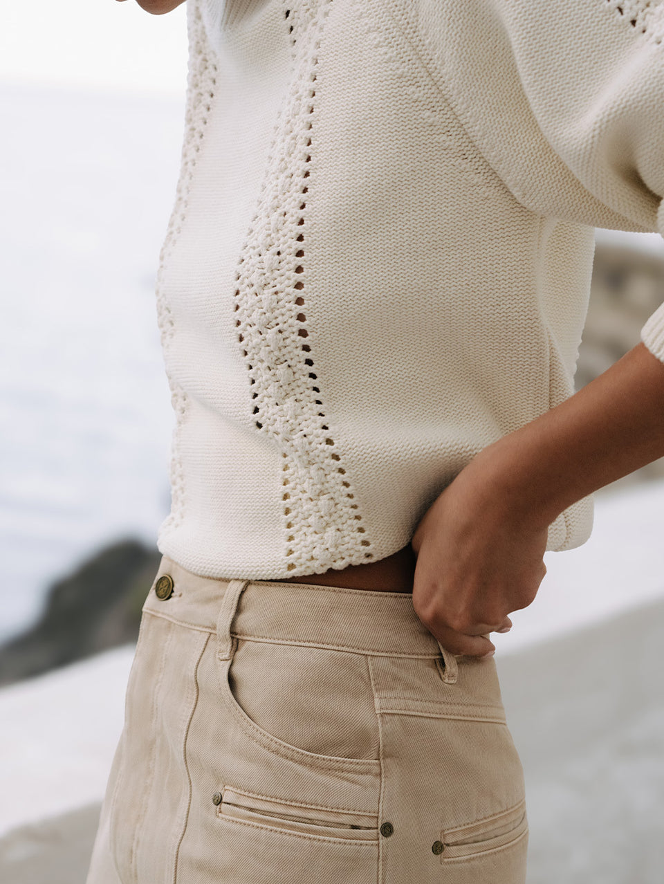 Close-up of a person wearing a white knit sweater and beige pants with a blurred background
