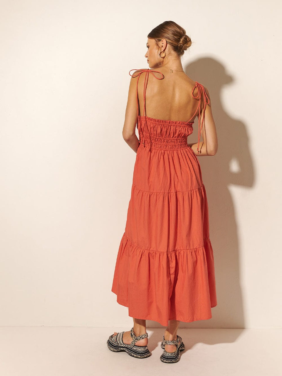 Back shot: Studio model wears KIVARI Casini dress - a coral block colour dress with thin straps, elasticated waist and tiered skirt.