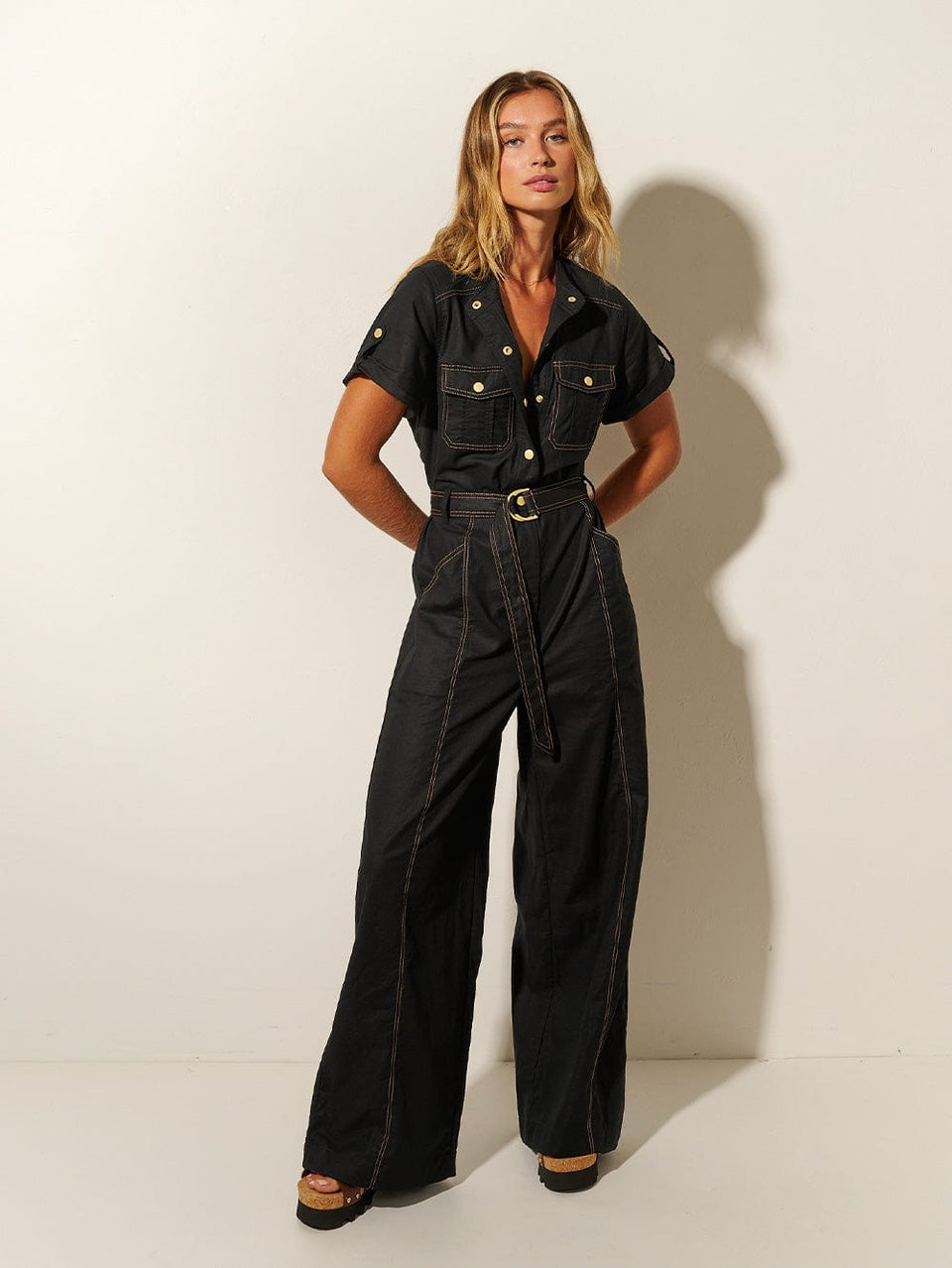 Studio model wears KIVARI Ebony Jumpsuit: a black linen jumpsuit with gold buttons, topstitch details, a button-front with signature hardware and belt.