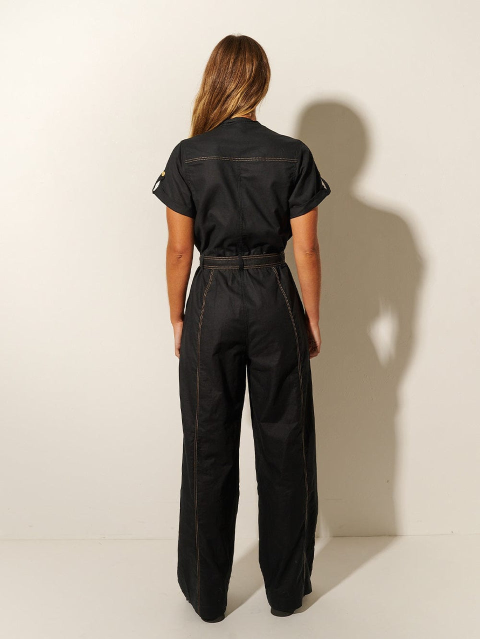 Back shot: Studio model wears KIVARI Ebony Jumpsuit: a black linen jumpsuit with gold buttons, topstitch details, a button-front with signature hardware and belt.