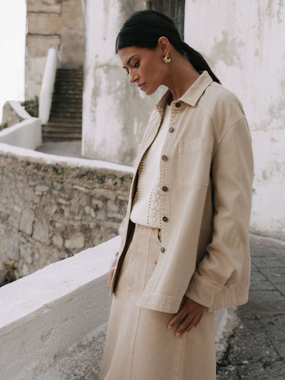 Woman wearing a beige jacket and pants standing against a stone wall.