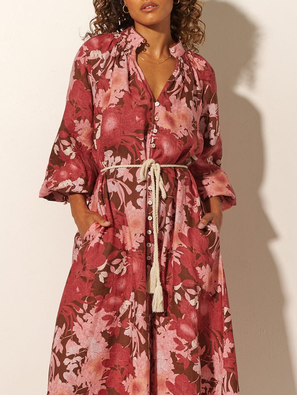 Close-up: Studio model wears KIVARI Hacienda Maxi Dress: A red, pink and brown floral dress with a shirred collar, button-through front, waist ties and full-length sleeves, crafted from sustainable LENZING Viscose Crepe.