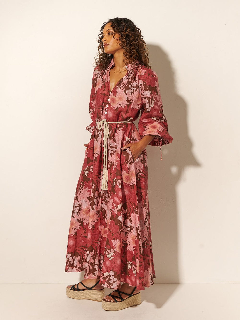 Close-up: Studio model wears KIVARI Hacienda Maxi Dress: A red, pink and brown floral dress with a shirred collar, button-through front, waist ties and full-length sleeves, crafted from sustainable LENZING Viscose Crepe.