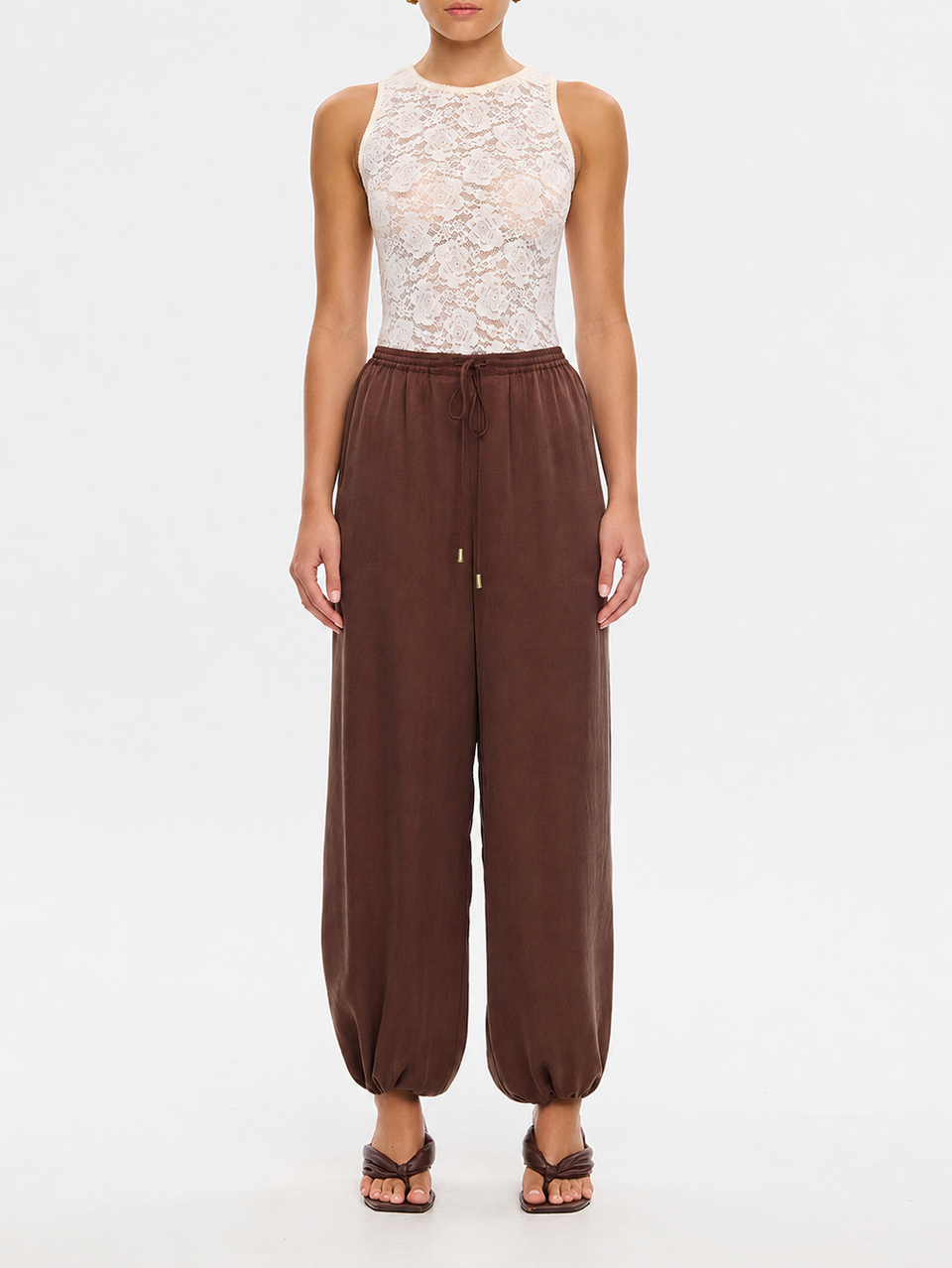 A model wearing a sleeveless white lace top and brown drawstring harem pants, standing against a plain white studio background.