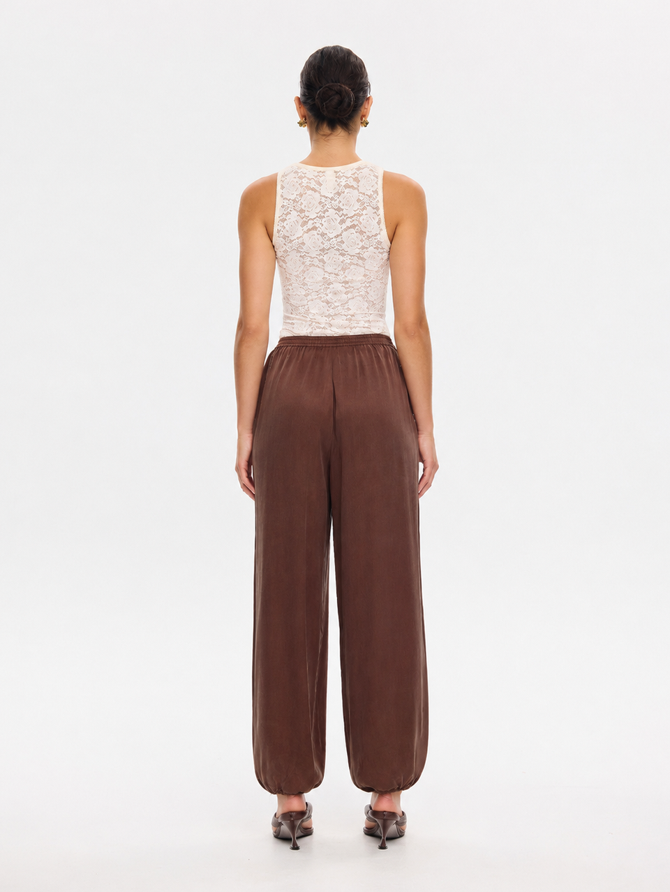 A model viewed from the back wearing a sleeveless cream lace top and brown wide-leg pants, standing against a plain white studio background.