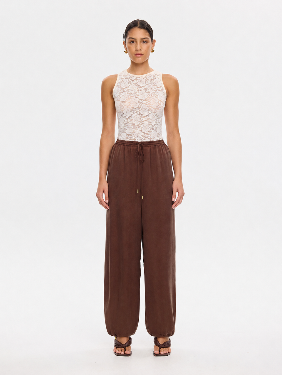 A model stands against a plain white studio background wearing a sleeveless white lace top and wide-leg brown pants with a drawstring waist.