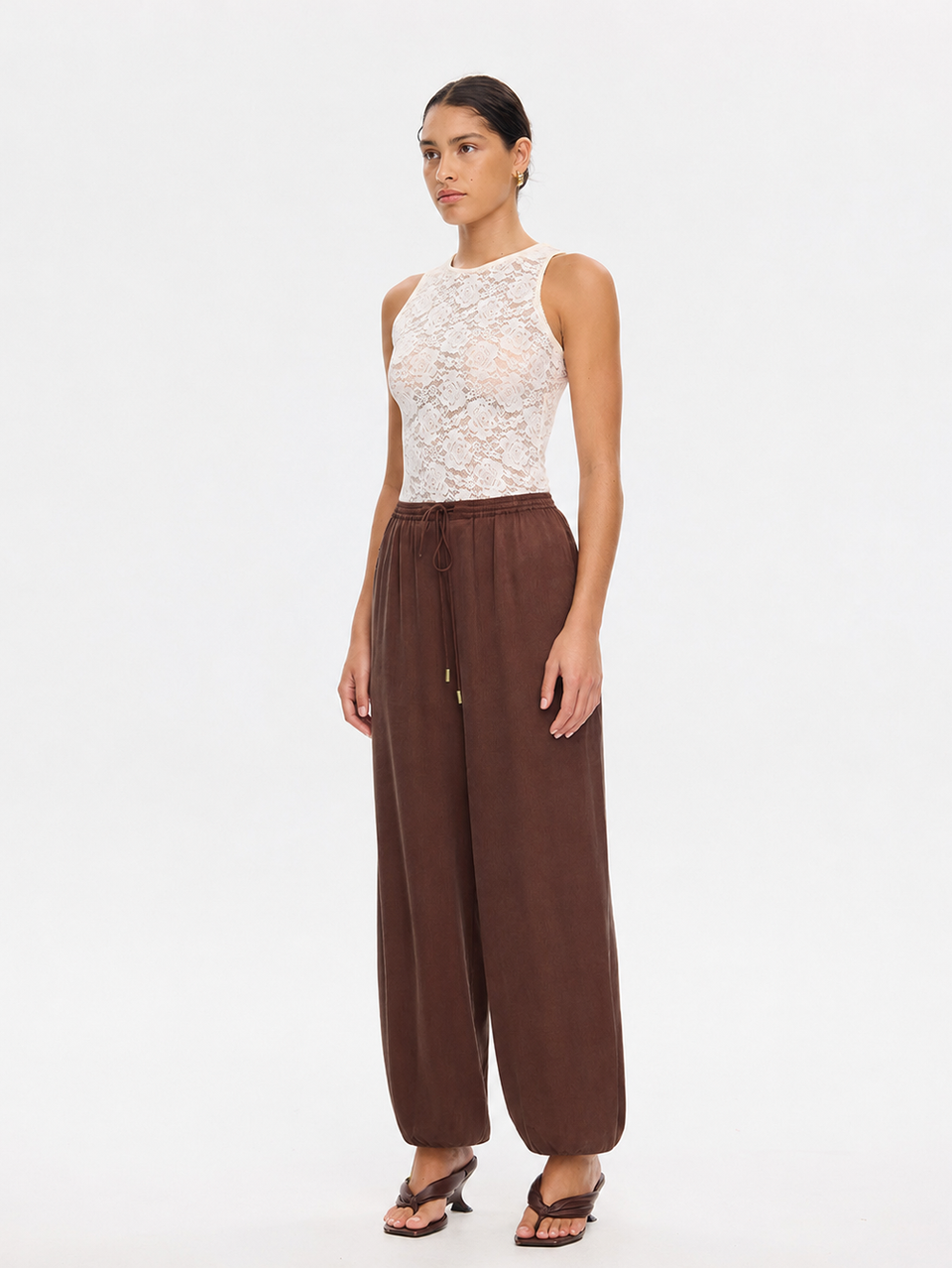 A model stands against a plain white studio background wearing a sleeveless white lace bodysuit top and wide-leg brown pants with sandals.