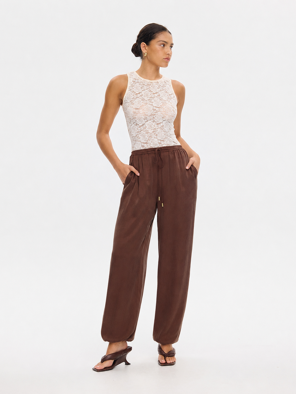 A woman modeling a sleeveless white lace top and brown wide-leg pants against a plain white studio background.