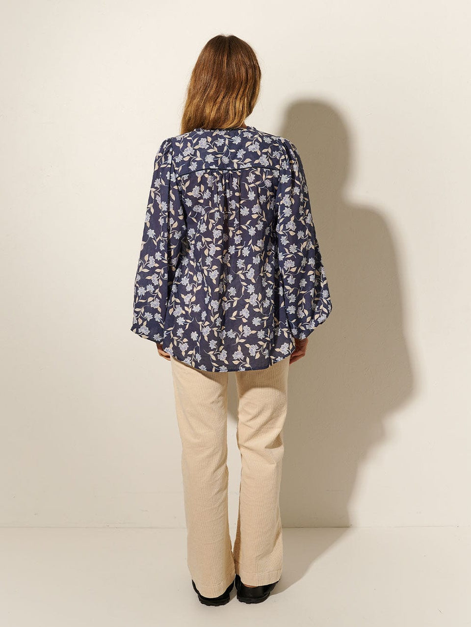 Back shot: Studio model wears KIVARI Jeanne Blouse: A navy and sky blue floral blouse made from cotton and featuring a button front, full-length blouson sleeves and frill collar detail.