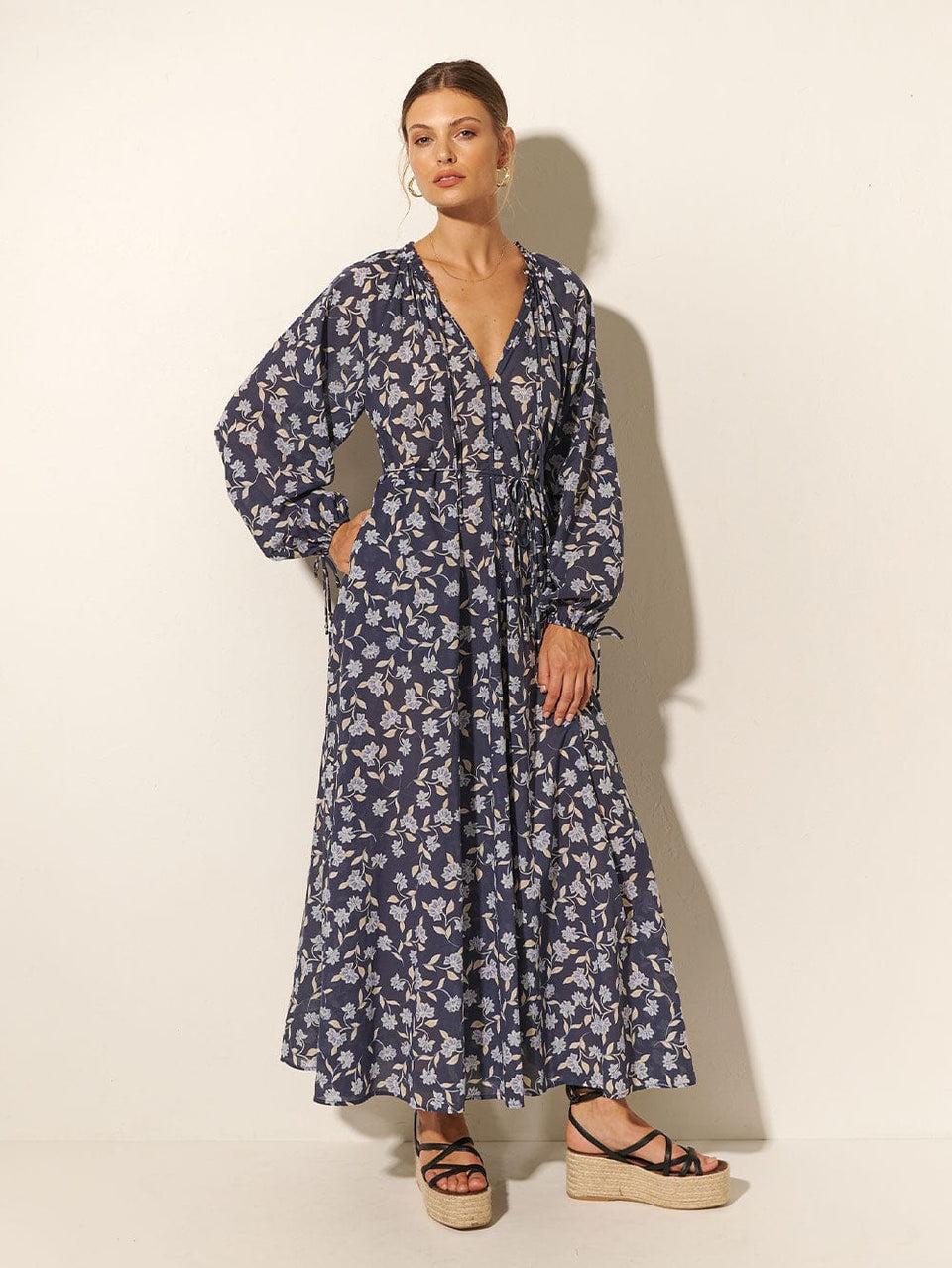 Studio model wears KIVARI Jeanne Maxi Dress: A navy and sky blue floral dress made from cotton and featuring a button front, waist tie, full-length blouson sleeves and frill collar detail.