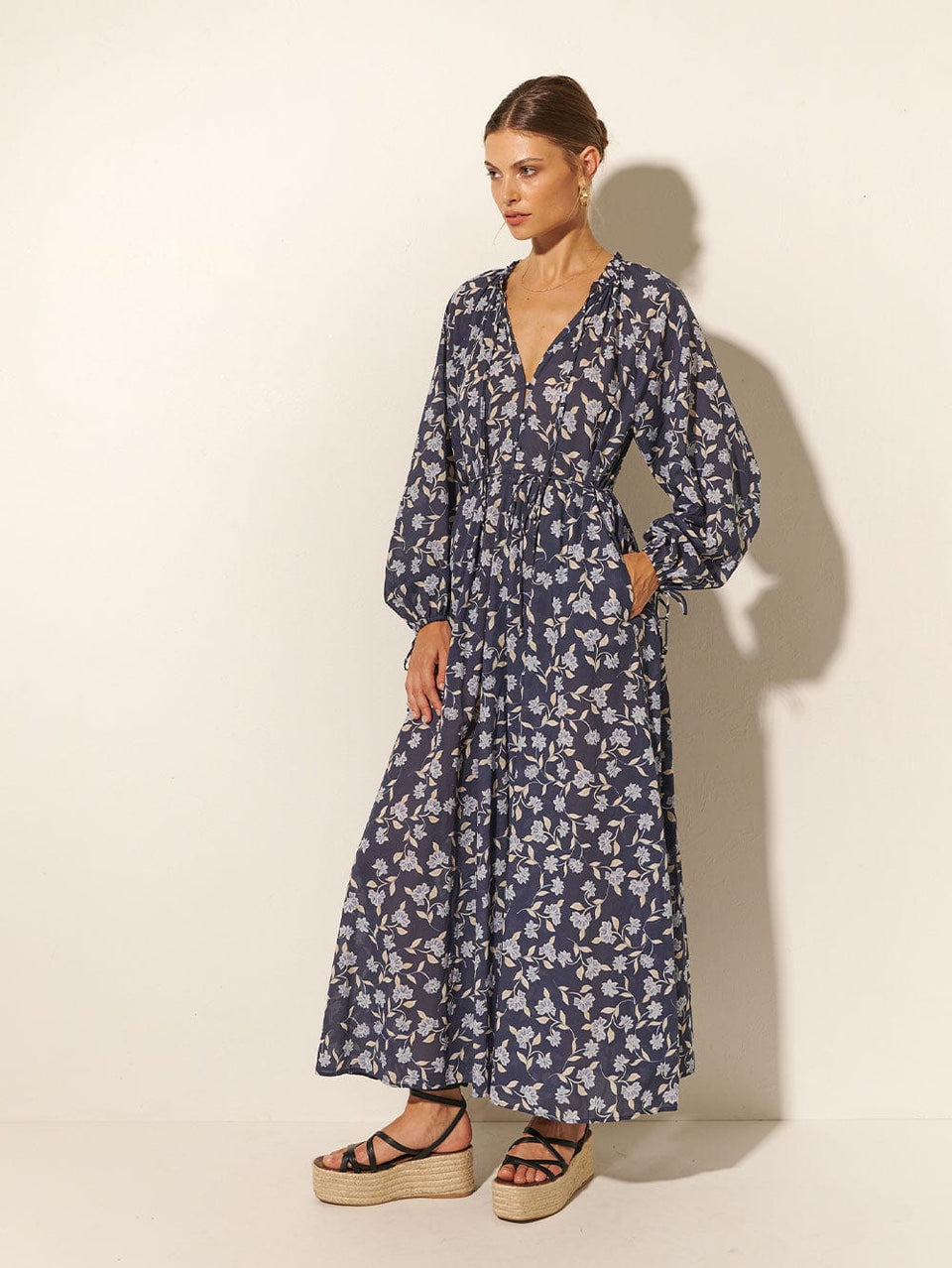Studio model wears KIVARI Jeanne Maxi Dress: A navy and sky blue floral dress made from cotton and featuring a button front, waist tie, full-length blouson sleeves and frill collar detail.