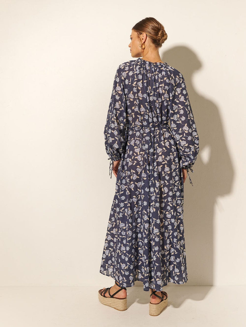 Back shot: Studio model wears KIVARI Jeanne Maxi Dress: A navy and sky blue floral dress made from cotton and featuring a button front, waist tie, full-length blouson sleeves and frill collar detail.