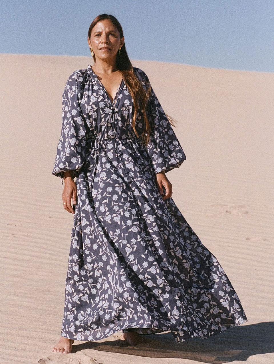 First Nations elder, Mary Pappin, in Mungo National Park wearing the KIVARI Jeanne Maxi Dress: A navy and sky blue floral dress made from cotton and featuring a button front, waist tie, full-length blouson sleeves and frill collar detail.