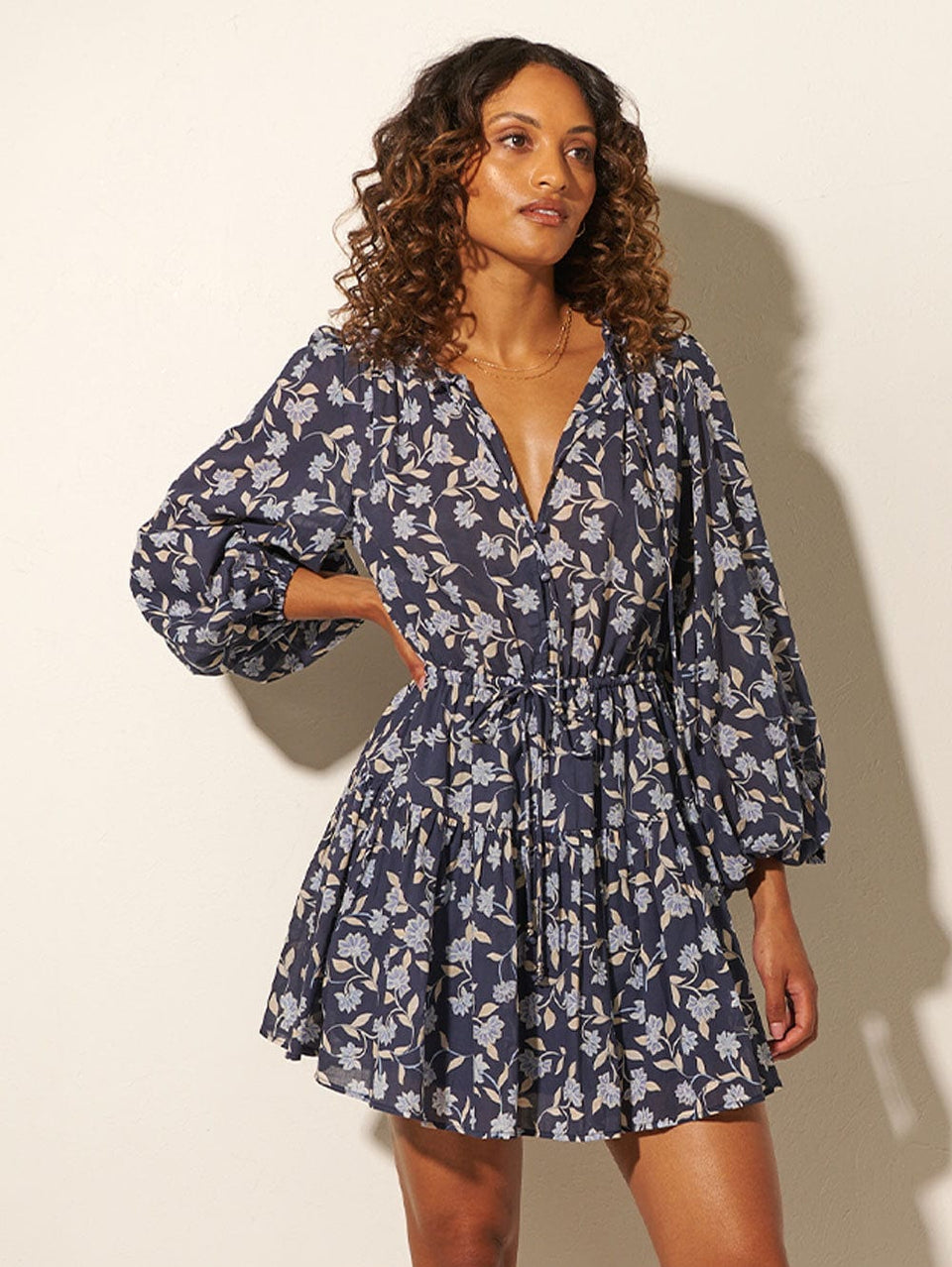 Studio model wears KIVARI Jeanne Mini Dress: A navy and sky blue floral dress made from cotton and featuring a button front, elasticated waist tie, full-length blouson sleeves and frill collar detail.