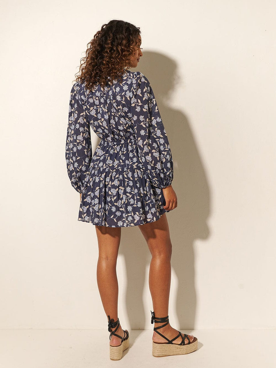Back shot: Studio model wears KIVARI Jeanne Mini Dress: A navy and sky blue floral dress made from cotton and featuring a button front, elasticated waist tie, full-length blouson sleeves and frill collar detail.