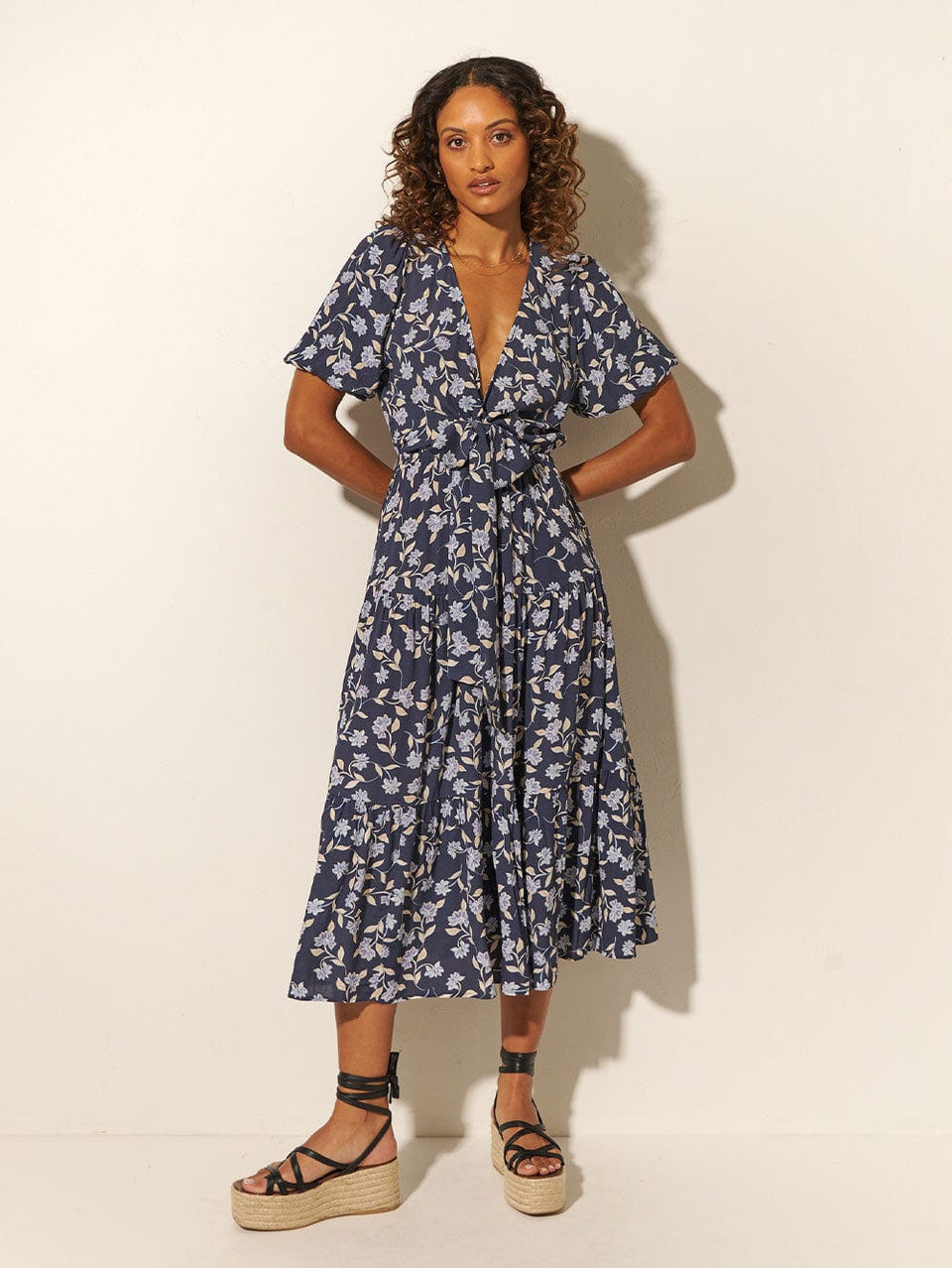 Studio model wears KIVARI Jeanne Tie Front Midi Dress: A navy and sky blue floral dress made from sustainable LENZING Viscose Crepe and featuring a tie front, elasticated waist, short puff sleeves and tiered skirt.