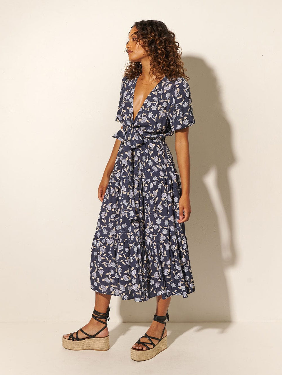 Studio model wears KIVARI Jeanne Tie Front Midi Dress: A navy and sky blue floral dress made from sustainable LENZING Viscose Crepe and featuring a tie front, elasticated waist, short puff sleeves and tiered skirt.