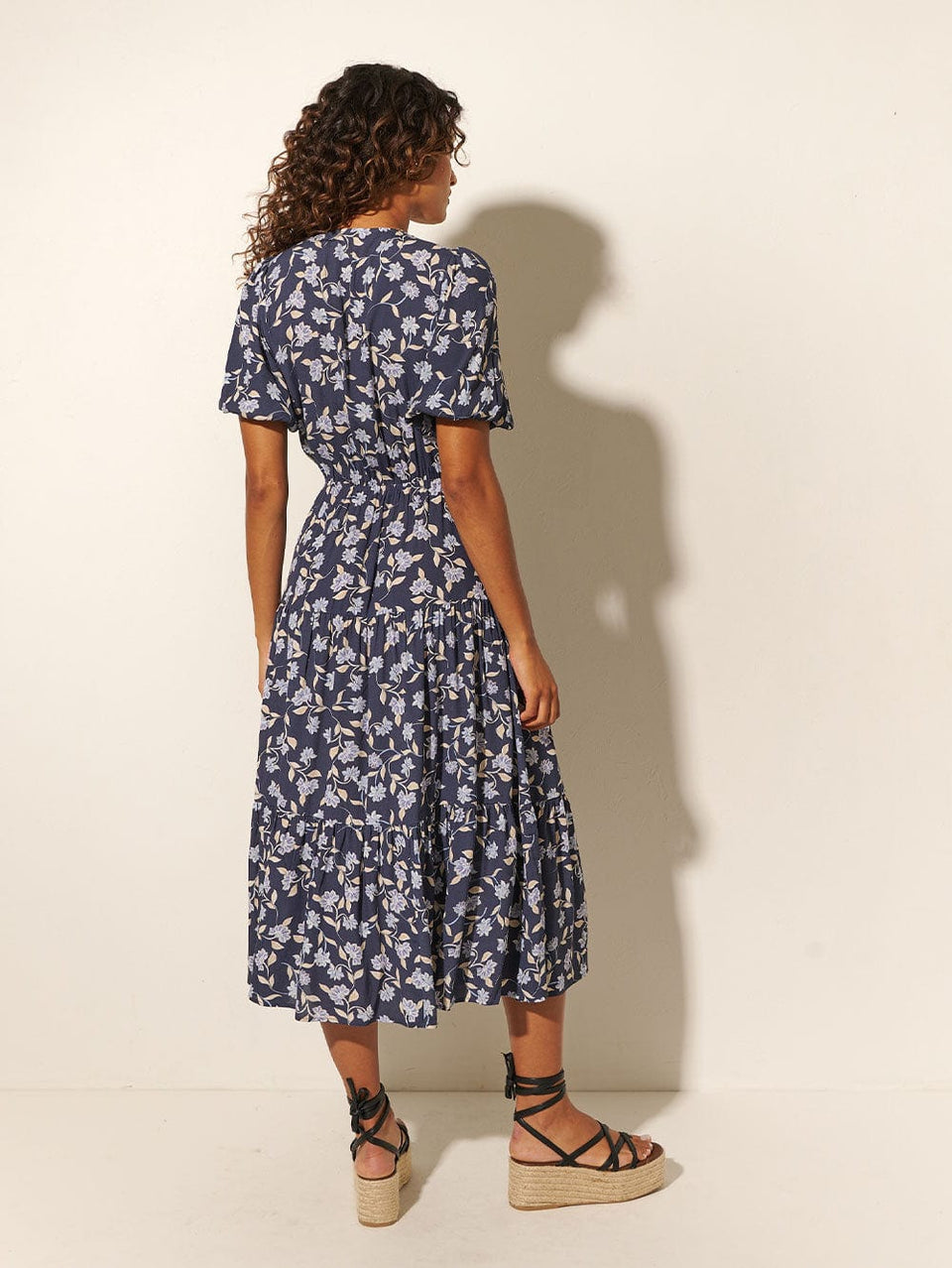 Back shot: Studio model wears KIVARI Jeanne Tie Front Midi Dress: A navy and sky blue floral dress made from sustainable LENZING Viscose Crepe and featuring a tie front, elasticated waist, short puff sleeves and tiered skirt.