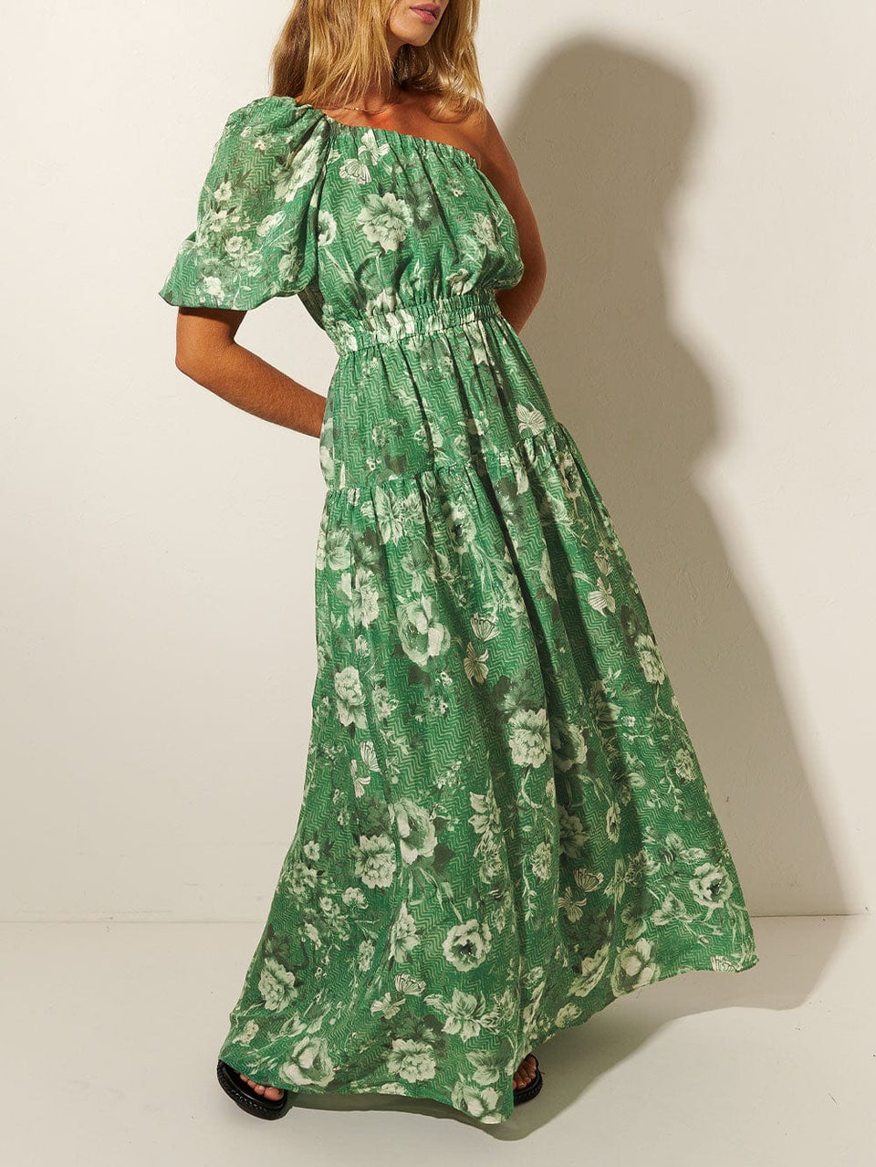 Studio model wears KIVARI Khalo Maxi Dress: A green floral one-shoulder dress with a short sleeve, elasticated front and waist, and tiered skirt.