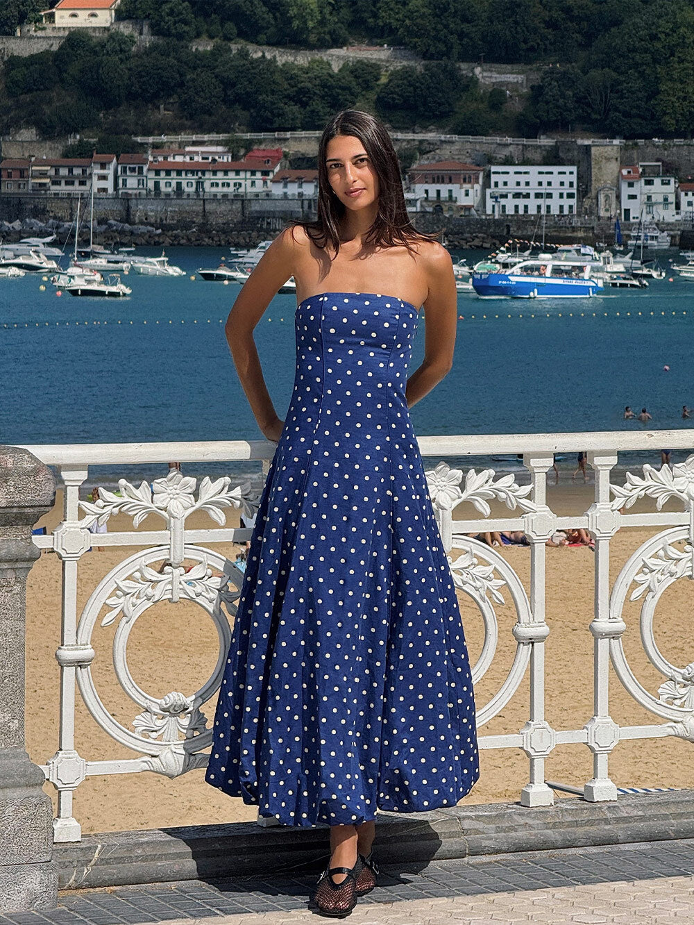 Woman in a blue polka dot dress standing by a waterfront with boats and buildings in the background.