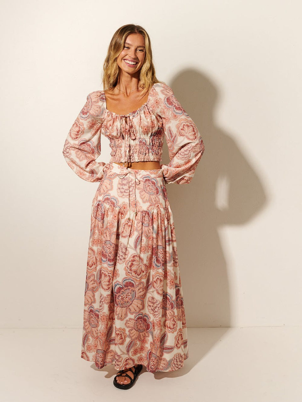 Studio model wears KIVARI Maya Crop Top: A pink and red floral on a natural base featuring an elasticated neckline, full-length blouson sleeves and a shirred bodice.