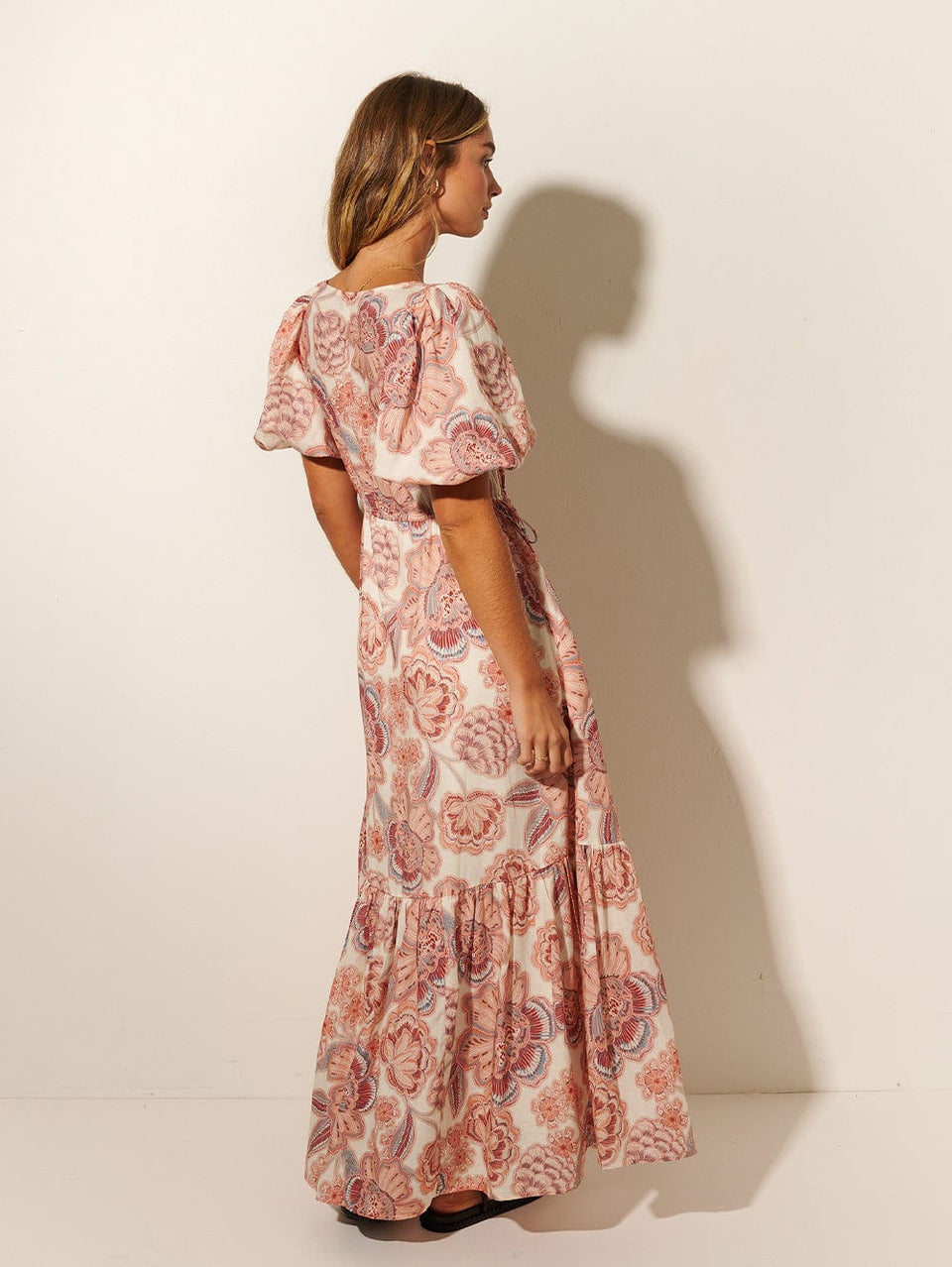 Back shot: Studio model wears the KIVARI Maya Maxi Skirt: A pink and red floral on a natural base featuring a voluminous puff sleeves, drawcord waist ties and a gathered tiered skirt with side pockets. 
