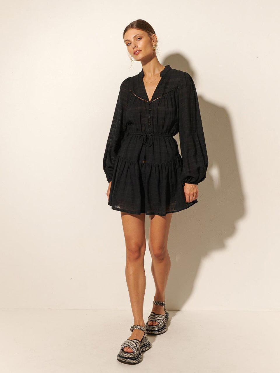 Studio model wears the KIVARI Rafaelle Mini Dress: a black dress made from cotton check featuring a button-front bodice, full-length blouson sleeves, drawstring waist and gathered hem frill.