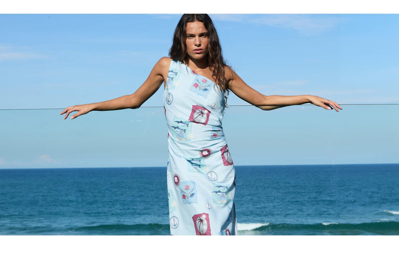 Woman in a patterned dress standing with arms outstretched against a blue sky and ocean.